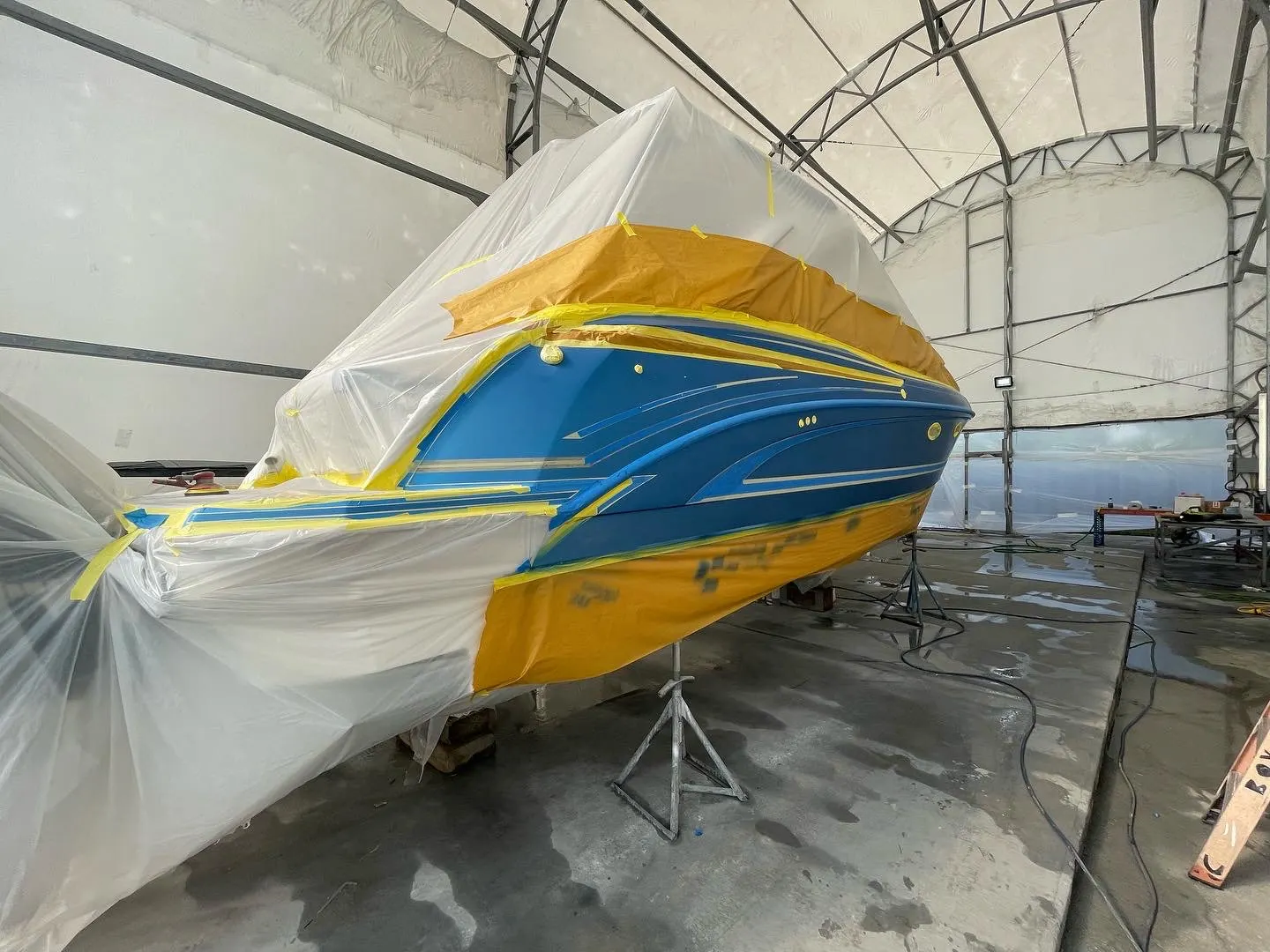 Blue powerboat hull side masked with plastic sheeting inside a marine work enclosure